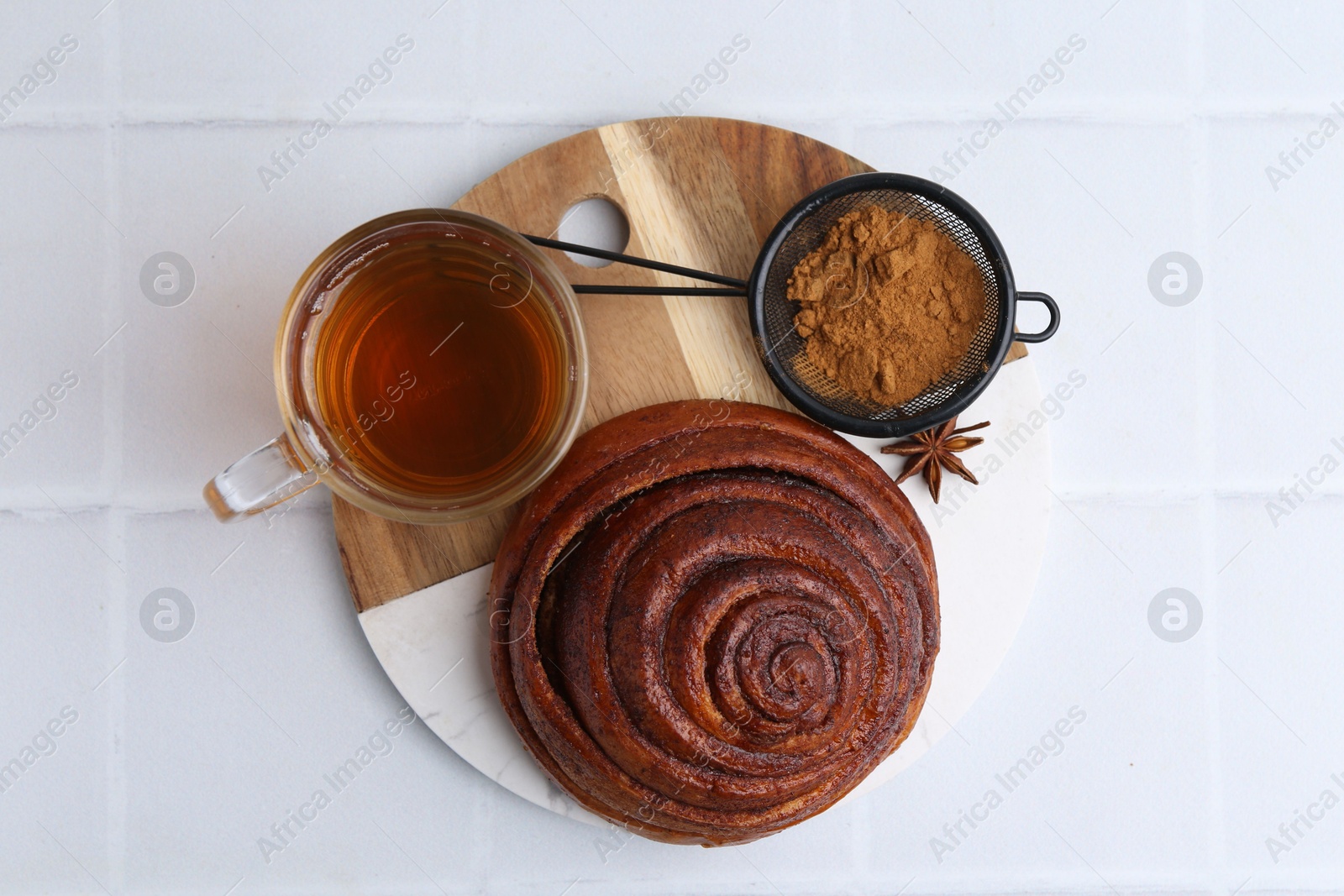 Photo of Delicious cinnamon roll bun, tea and spices on white tiled table, top view