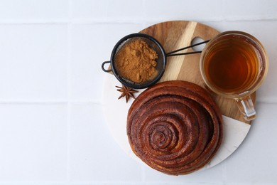 Photo of Delicious cinnamon roll bun, tea and spices on white tiled table, top view. Space for text