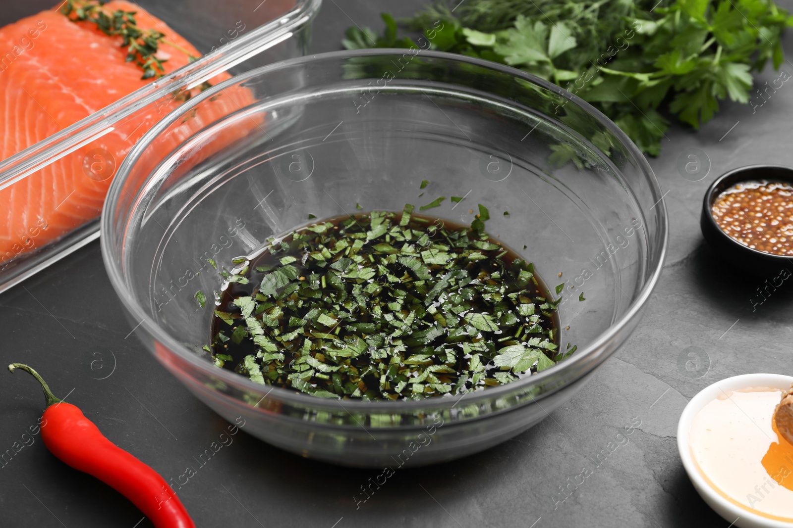 Photo of Soy marinade in bowl, salmon fillet and other products on dark gray textured table, closeup