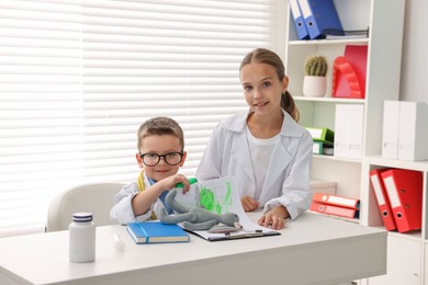 Photo of Little boy and girl pretending to be doctors at white table indoors