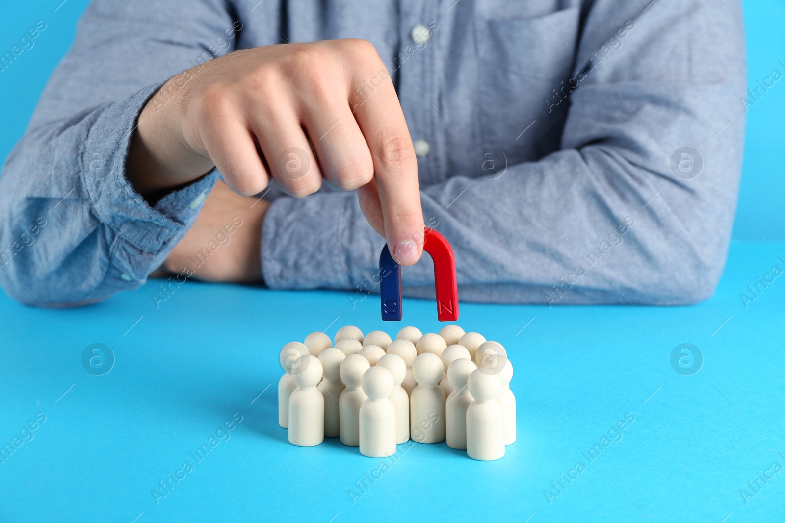 Photo of Man with magnet attracting wooden human figures on light blue background, closeup