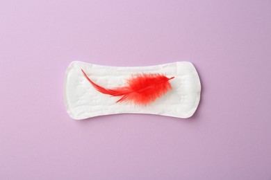 Photo of Menstrual pad with red feather on lilac background, top view