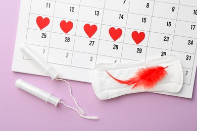 Photo of Calendar with marked dates, menstrual pad with feather and tampons on lilac background, top view