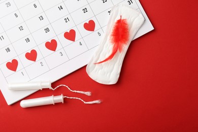 Photo of Calendar with marked dates, menstrual pad with feather and tampons on red background, top view