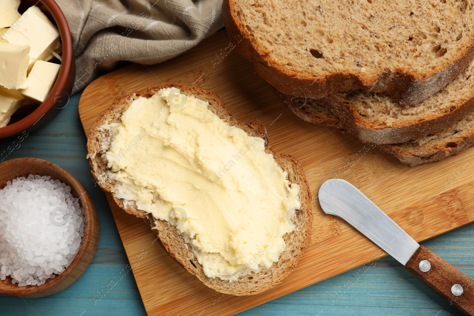 Photo of Fresh bread with butter, salt and knife on blue wooden table, flat lay