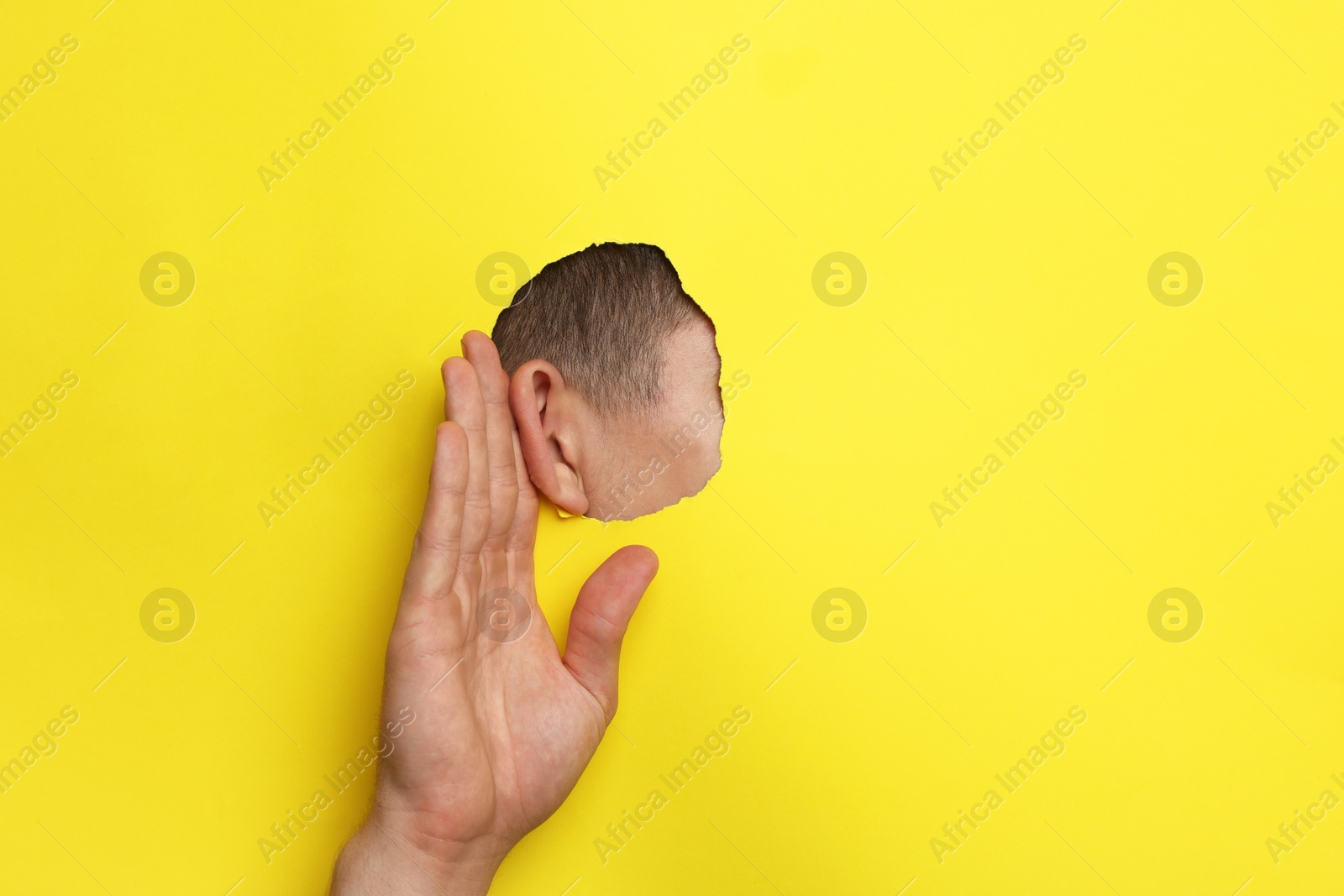 Photo of Man showing hand to ear gesture through hole in yellow paper, closeup