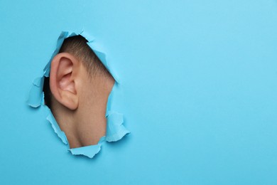 Man showing his ear through hole in light blue paper, closeup. Space for text