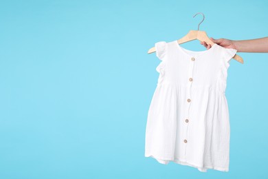 Photo of Woman holding hanger with white baby dress on light blue background, closeup. Space for text