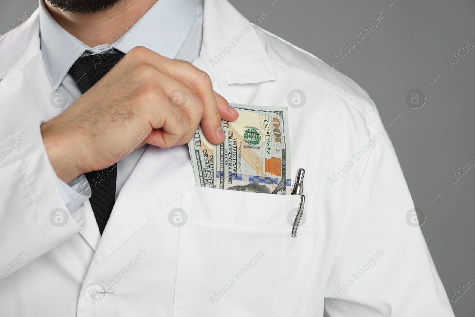 Photo of Corruption concept. Doctor putting dollar banknotes into his pocket on grey background, closeup