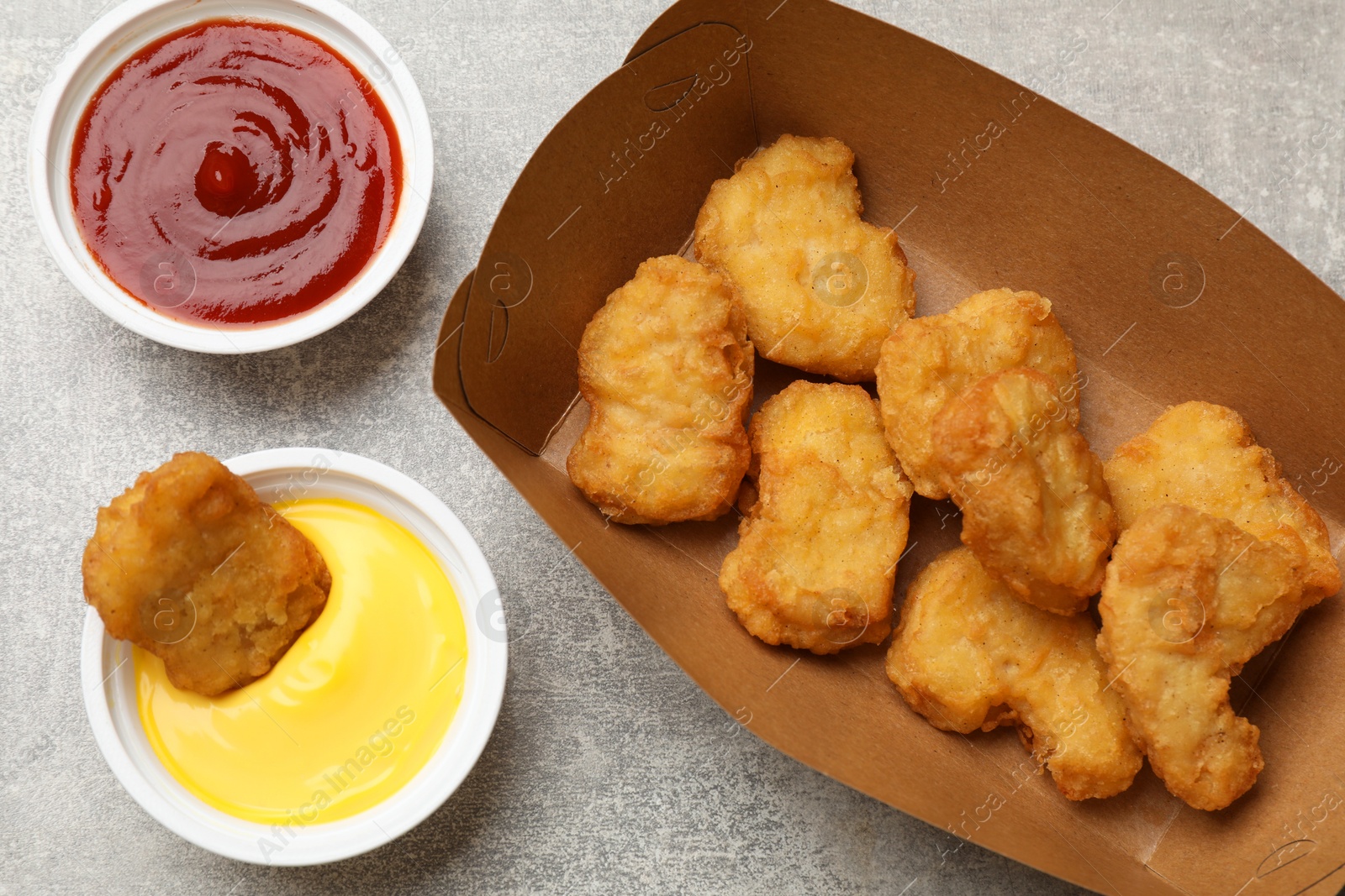 Photo of Delicious chicken nuggets in carton box and sauces on gray textured table, flat lay