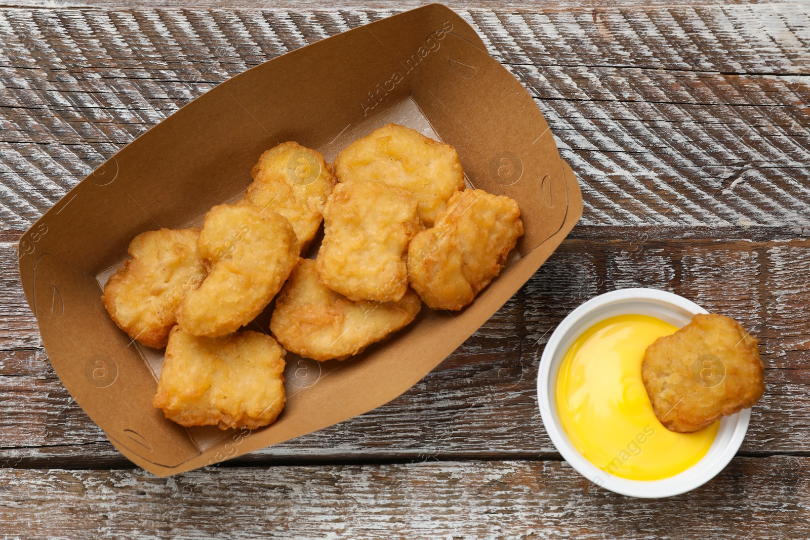 Photo of Delicious chicken nuggets in carton box and sauce on wooden table, flat lay