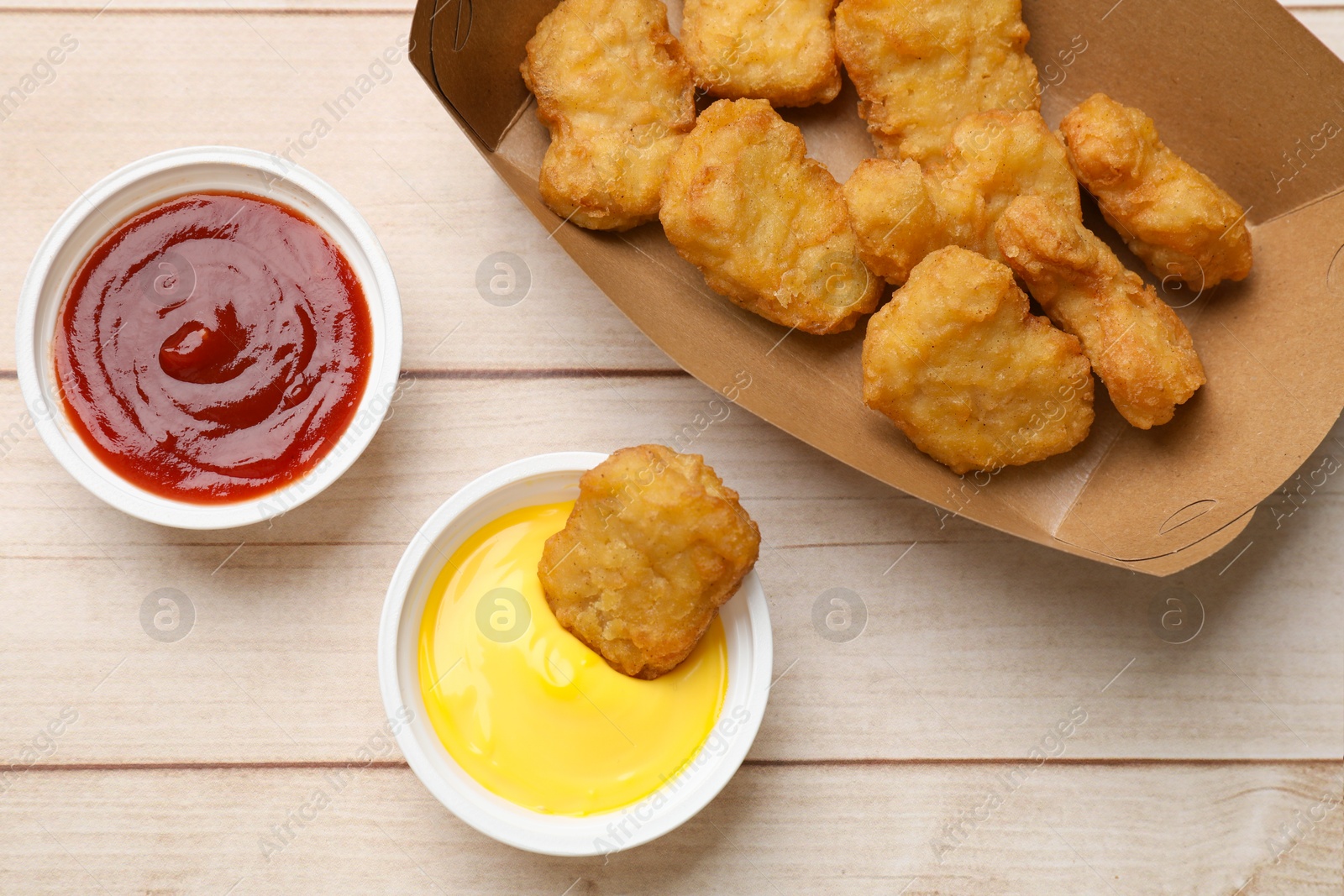 Photo of Delicious chicken nuggets in carton box and sauces on wooden table, flat lay