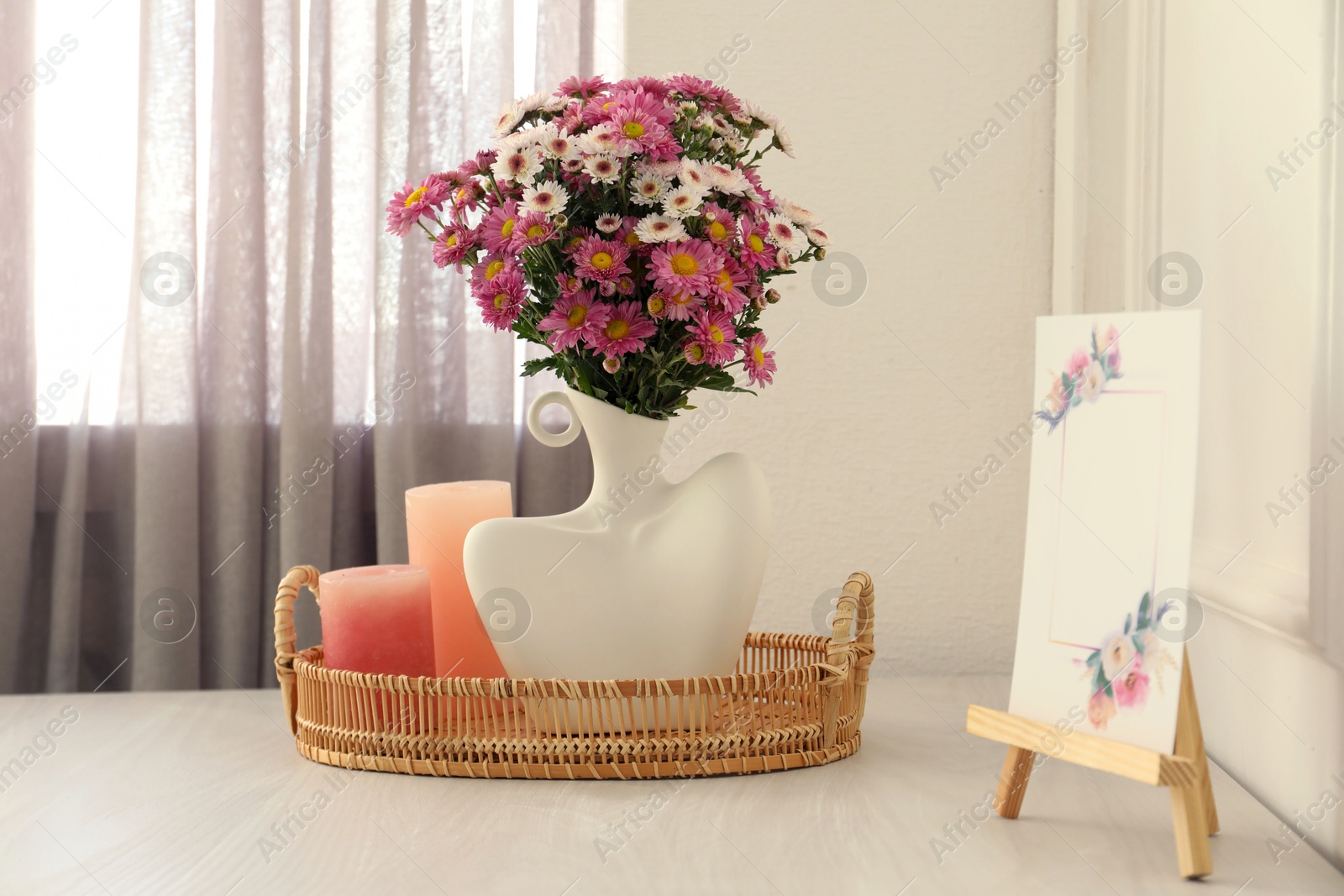 Photo of Stylish vase with beautiful flowers, candles and small easel on white wooden table at home