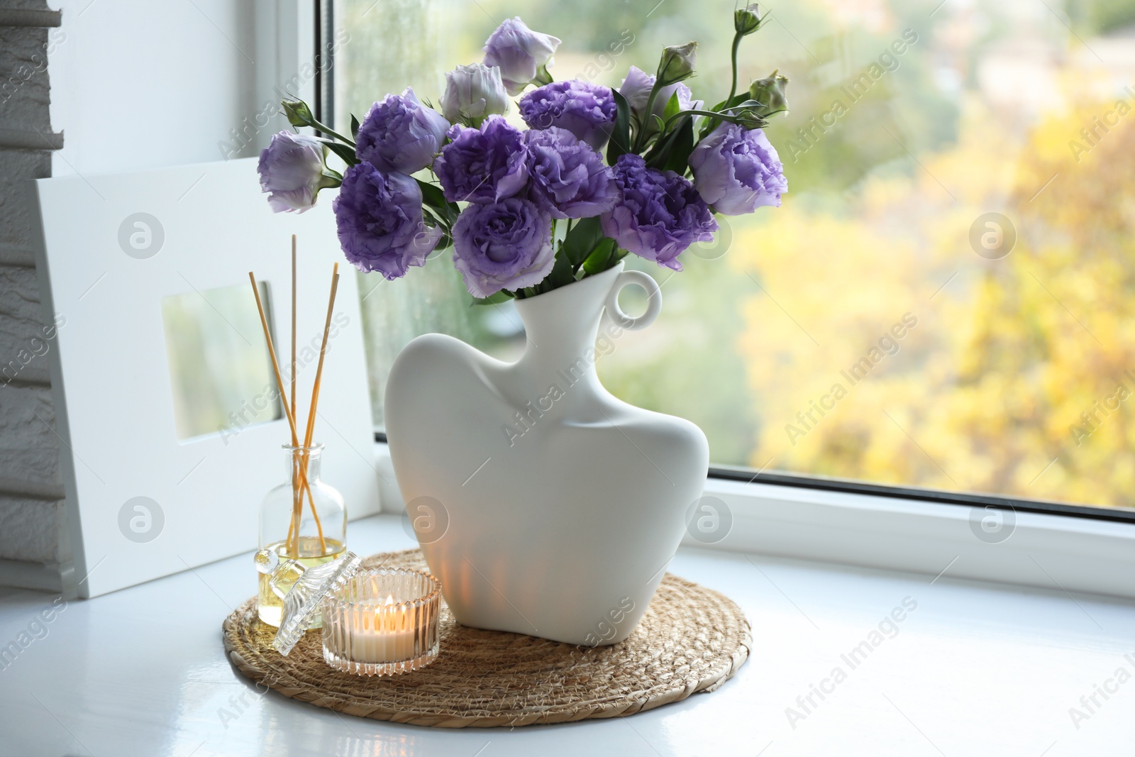 Photo of Stylish vase with beautiful flowers, candle and reed diffuser near window at home