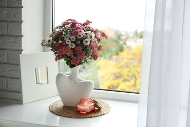 Photo of Stylish vase with beautiful flowers and burning candle near window at home