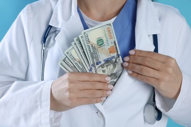 Photo of Corruption concept. Doctor putting dollar banknotes into her coat on light blue background, closeup