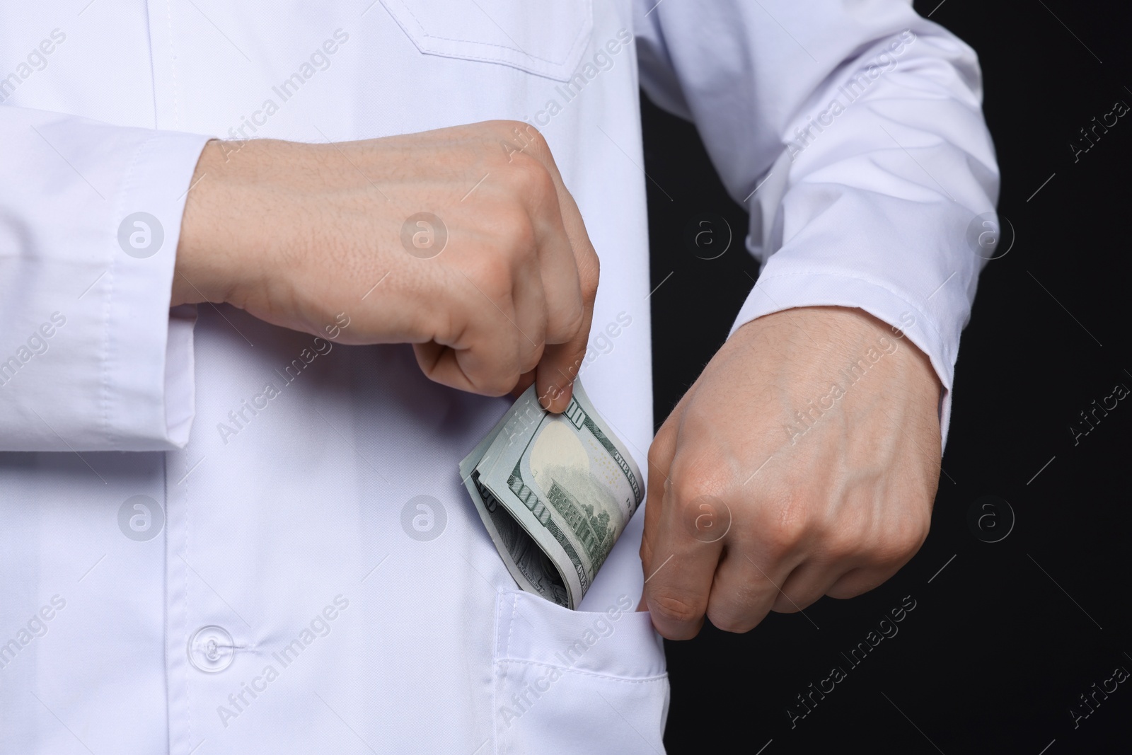 Photo of Corruption concept. Doctor putting dollar banknotes into his pocket on black background, closeup