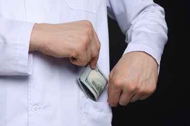 Photo of Corruption concept. Doctor putting dollar banknotes into his pocket on black background, closeup