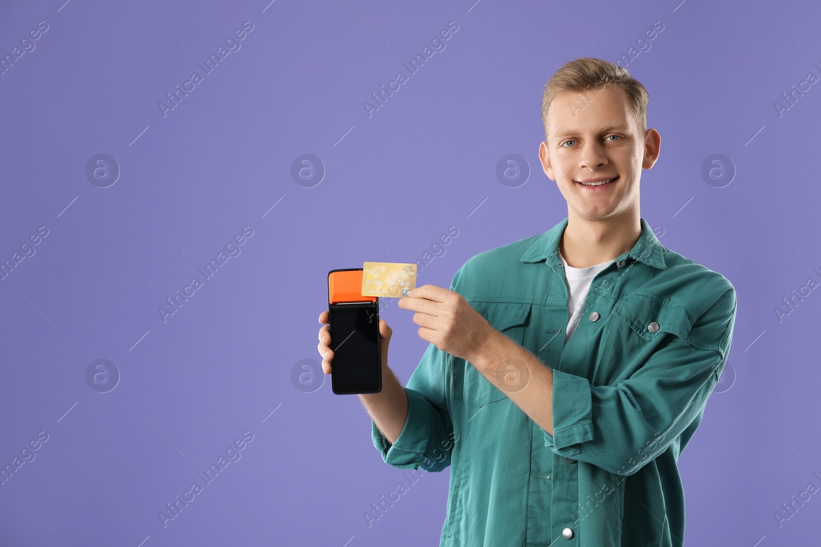 Photo of Happy young man with payment terminal and debit card on purple background, space for text