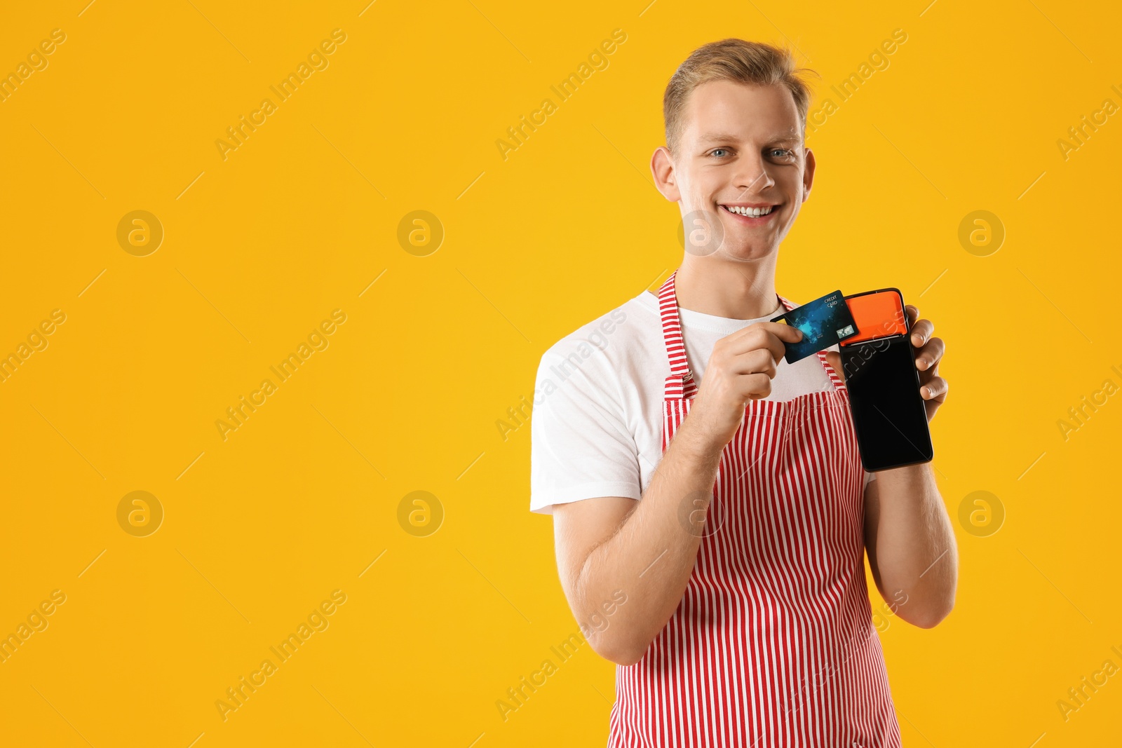 Photo of Happy young man in apron with payment terminal and debit card on yellow background, space for text