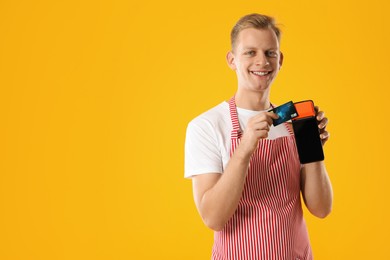 Photo of Happy young man in apron with payment terminal and debit card on yellow background, space for text