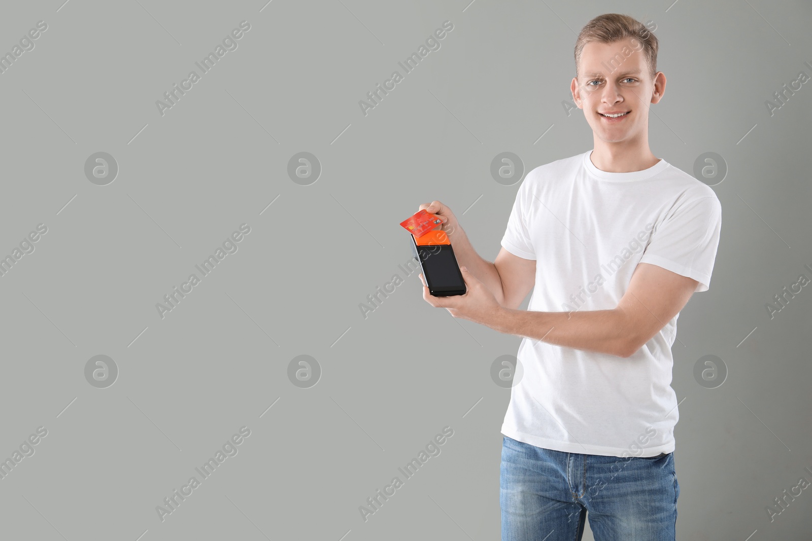 Photo of Happy young man with payment terminal and debit card on light grey background, space for text