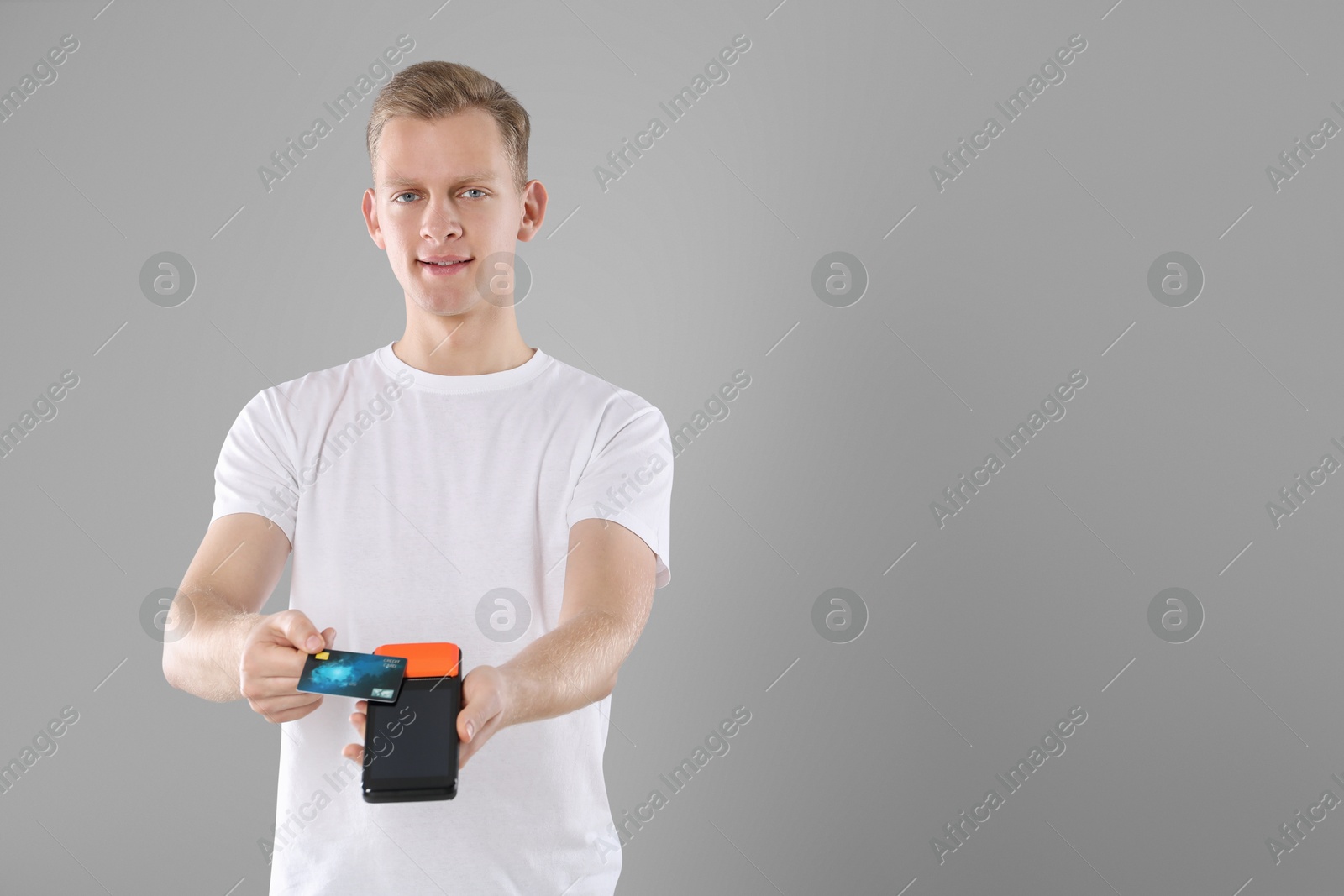 Photo of Happy young man with payment terminal and debit card on light grey background, space for text