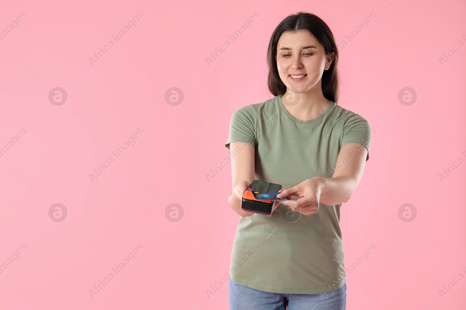 Photo of Happy young woman with payment terminal and debit card on pink background, space for text