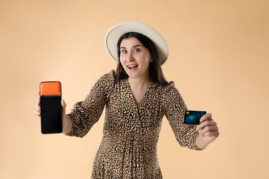 Photo of Happy young woman with payment terminal and debit card on beige background