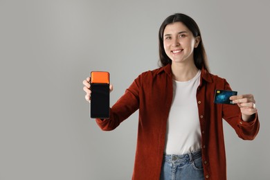 Photo of Happy young woman with payment terminal and debit card on gray background, space for text