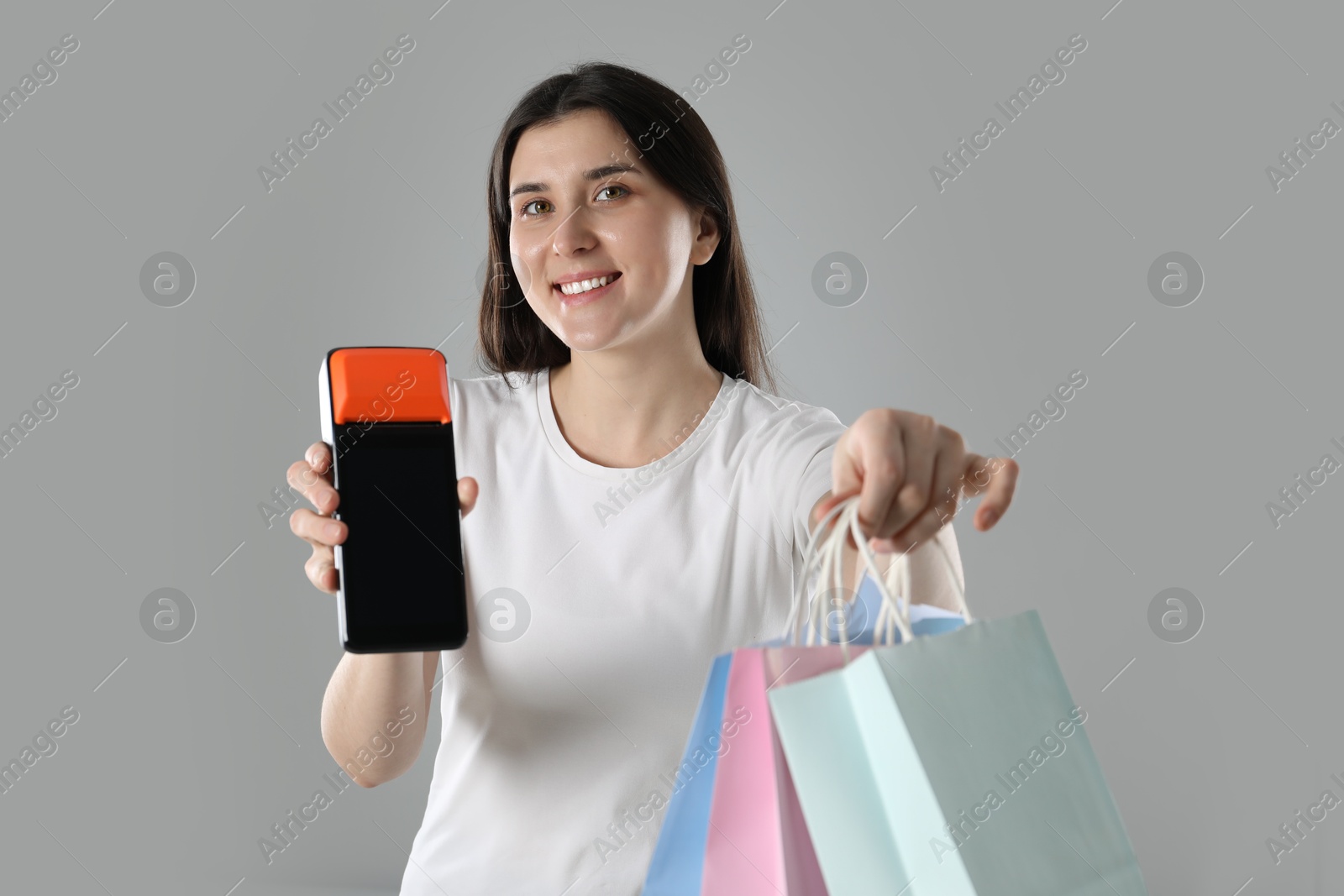 Photo of Happy young woman with payment terminal and shopping bags on gray background