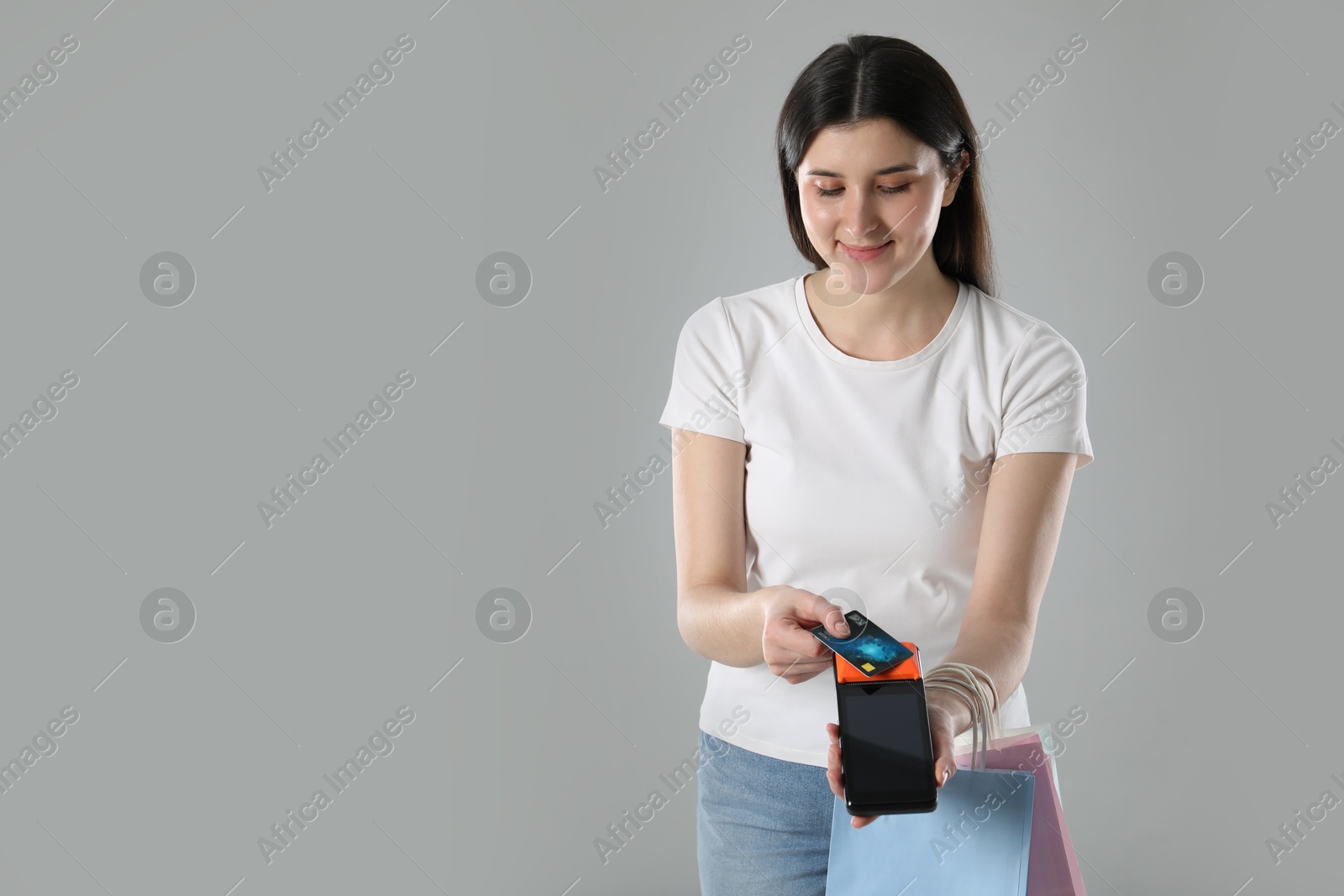 Photo of Young woman with payment terminal, shopping bags and debit card on gray background, space for text