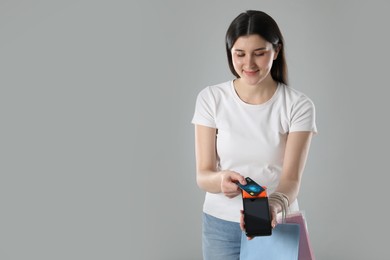 Young woman with payment terminal, shopping bags and debit card on gray background, space for text