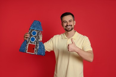 Photo of Happy man with rocket shaped pinata showing thumbs up on red background