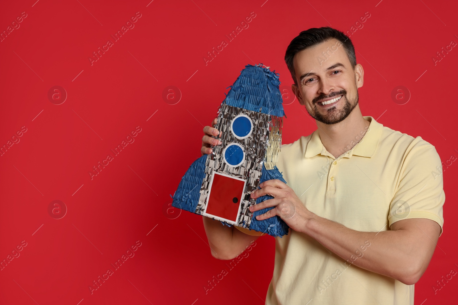 Photo of Happy man with rocket shaped pinata on red background, space for text