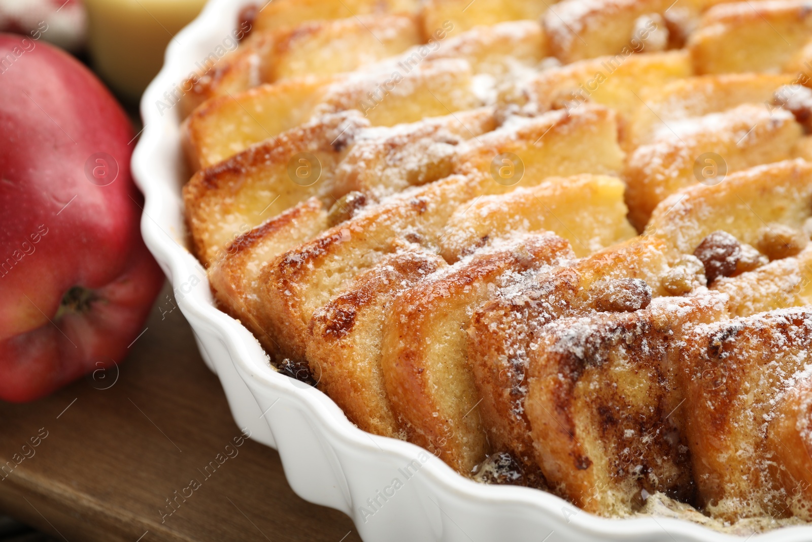 Photo of Delicious bread pudding with raisins, powdered sugar and apple on wooden table, closeup