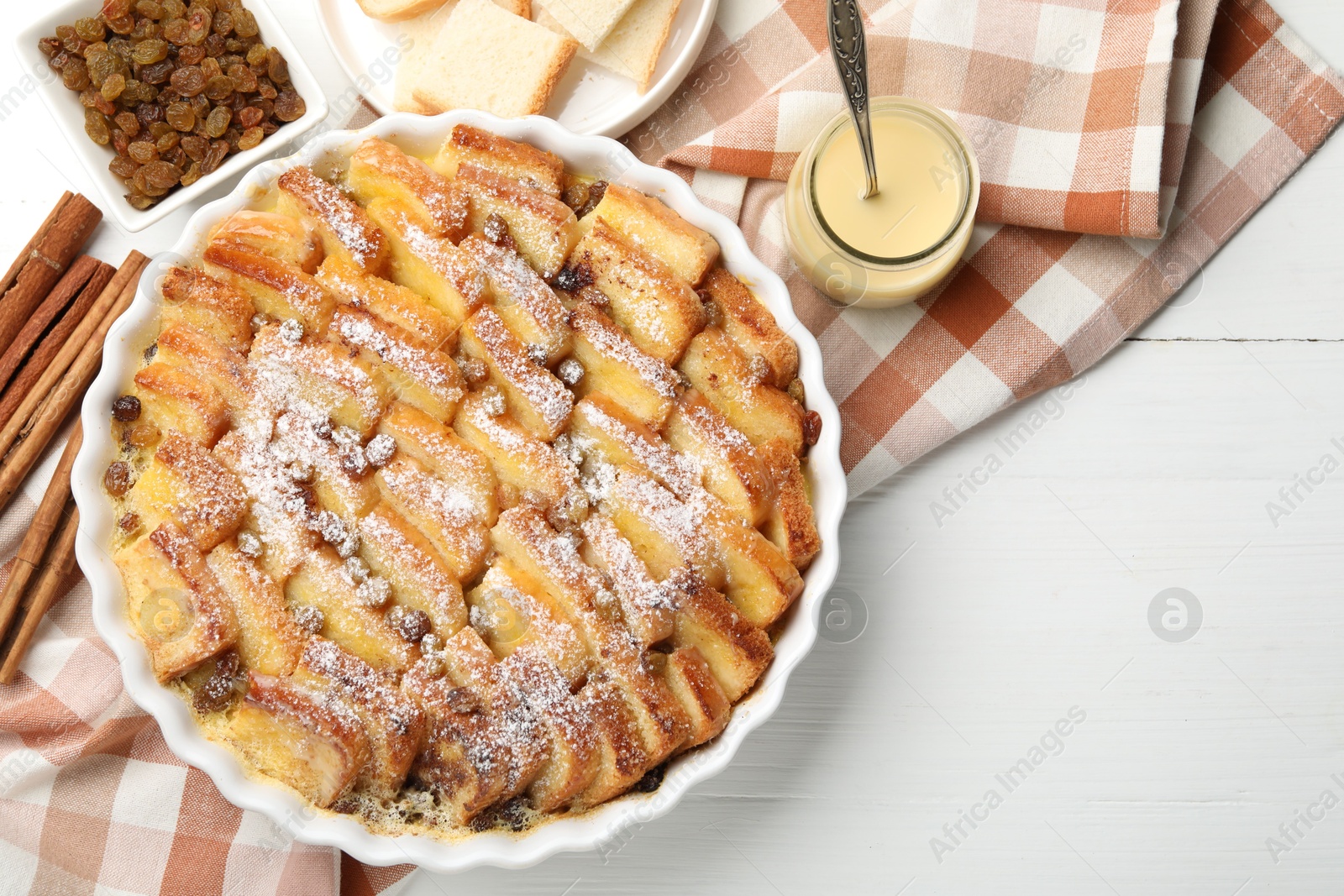 Photo of Delicious bread pudding with raisins, powdered sugar, condensed milk on white wooden table, flat lay