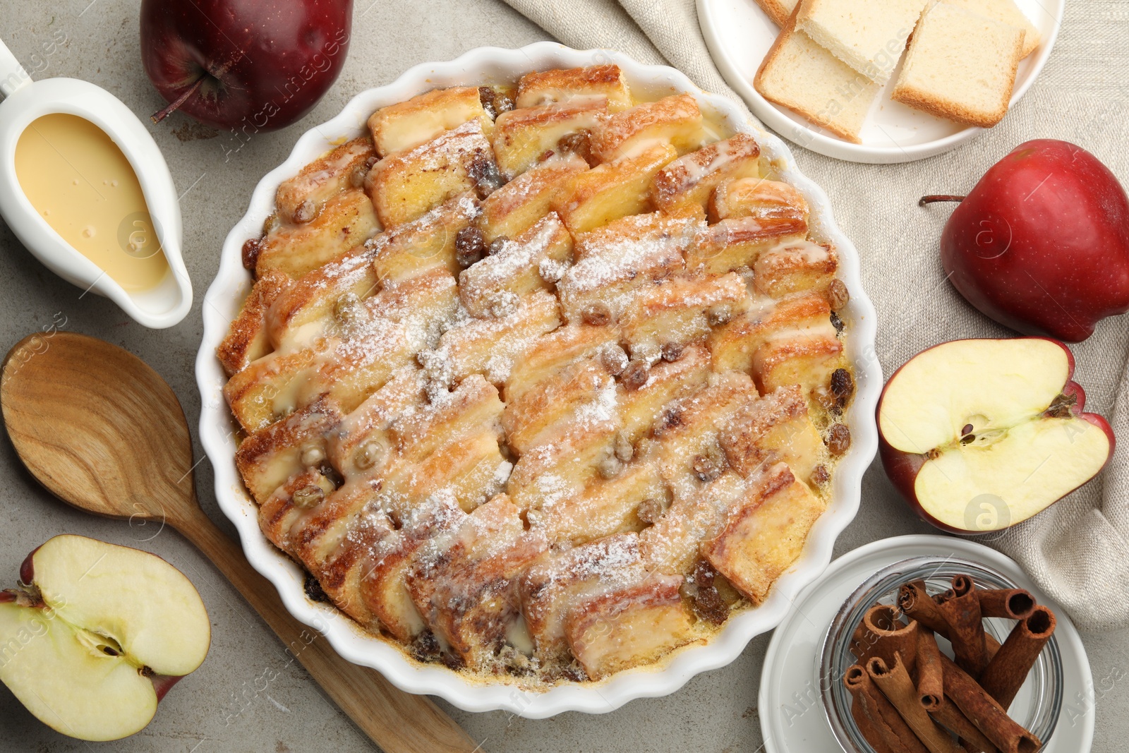 Photo of Delicious bread pudding with raisins, powdered sugar, cinnamon and apples on grey table, flat lay