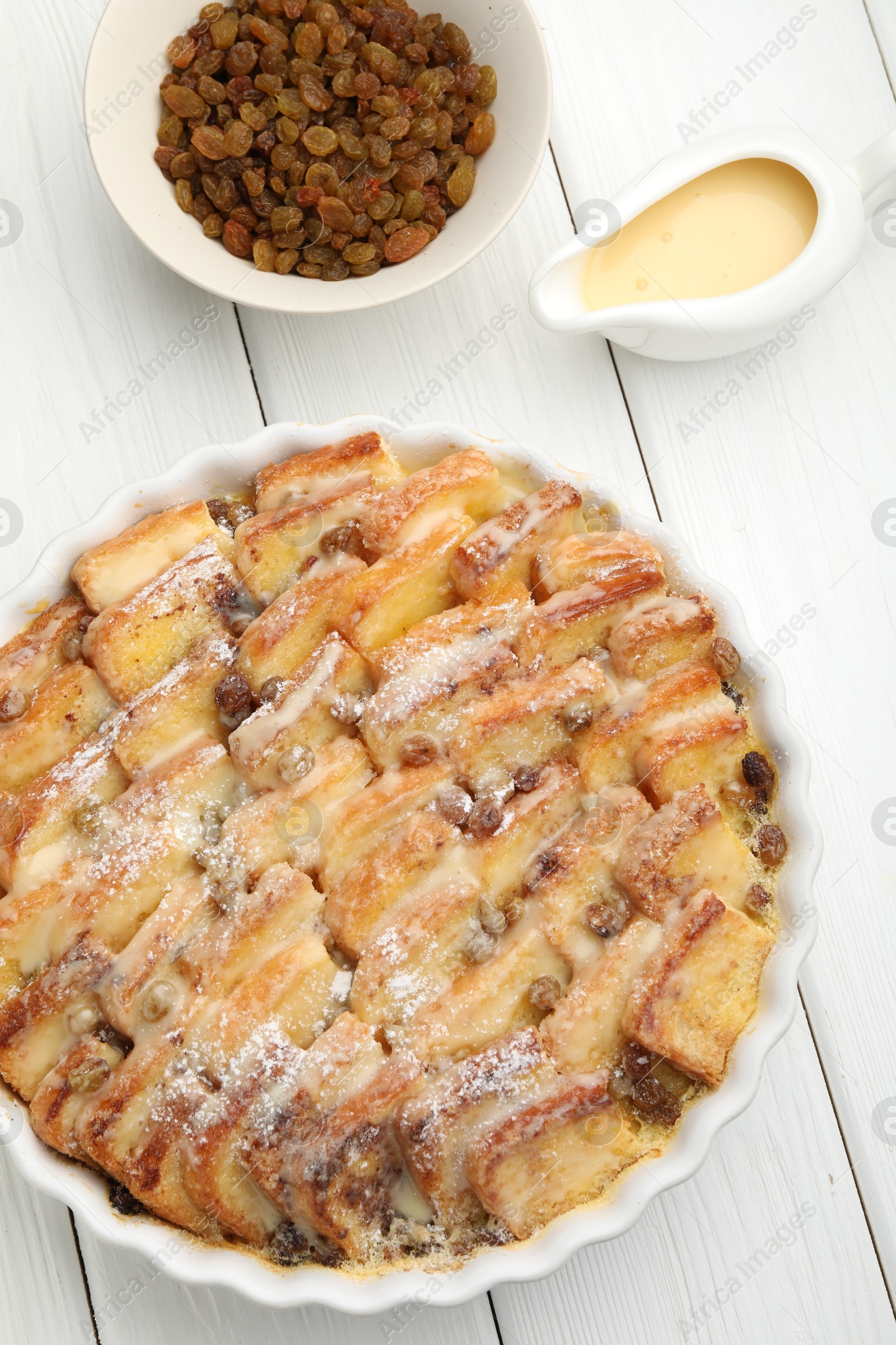 Photo of Delicious bread pudding with raisins, powdered sugar, condensed milk on white wooden table, flat lay