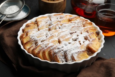 Photo of Delicious bread pudding with raisins, powdered sugar and tea on black table, closeup