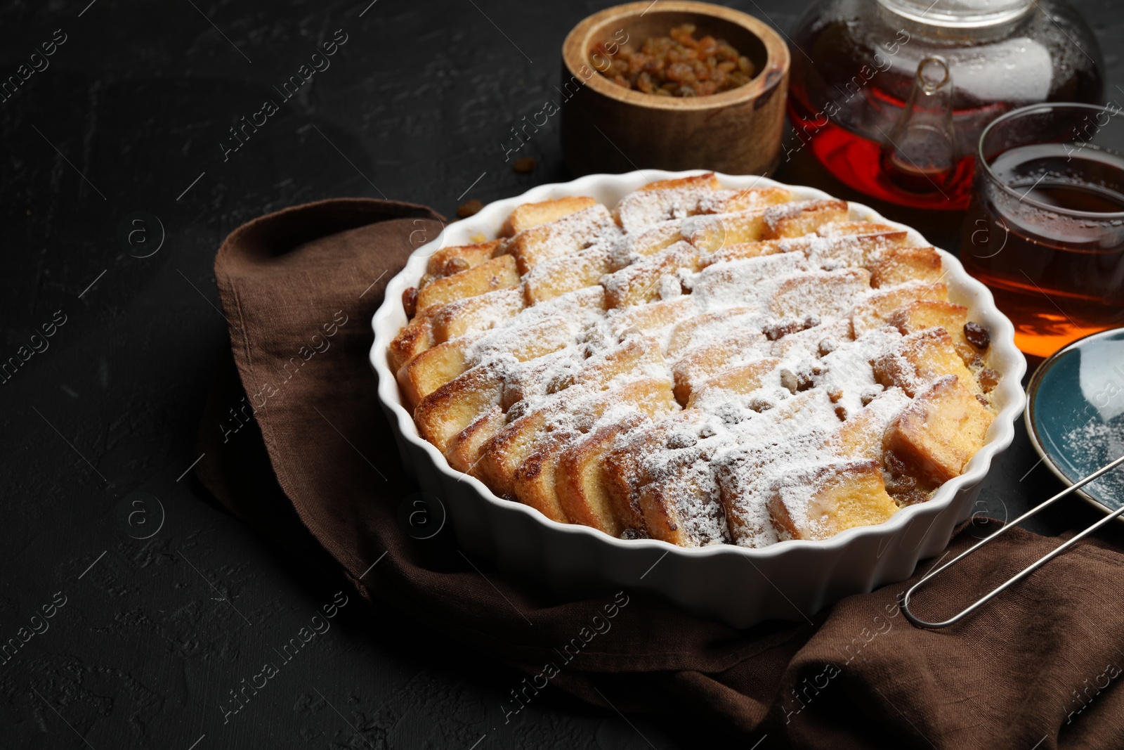 Photo of Delicious bread pudding with raisins, powdered sugar and tea on black table