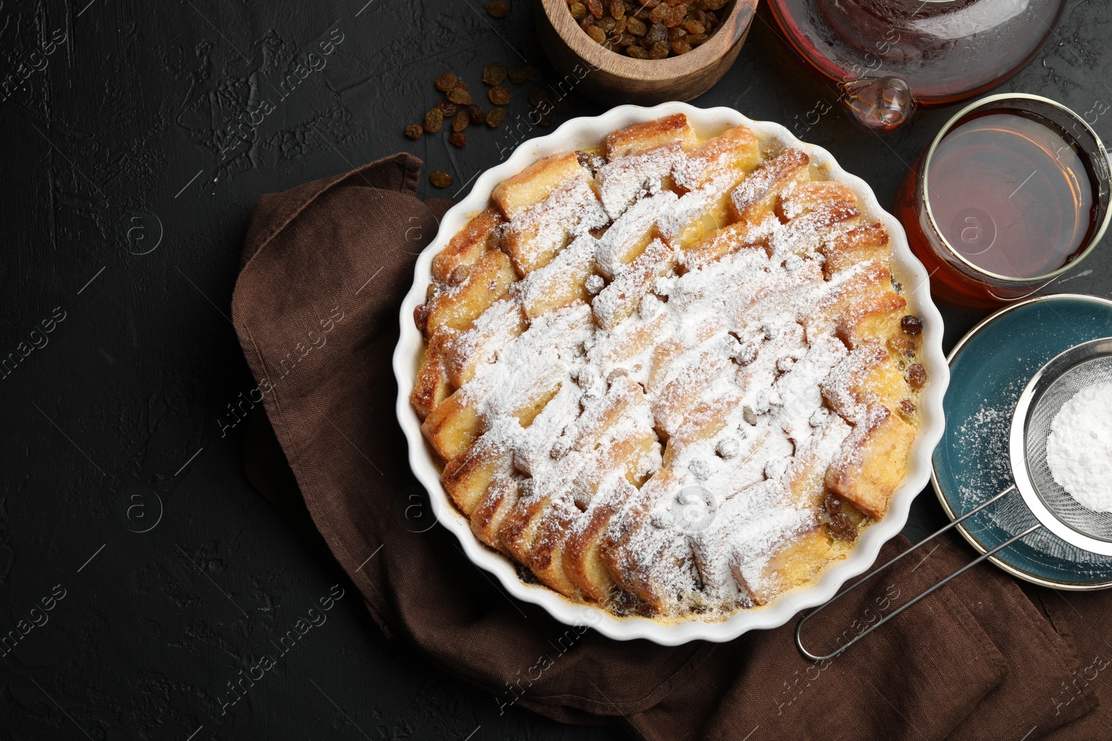 Photo of Delicious bread pudding with raisins, powdered sugar and tea on black table, flat lay