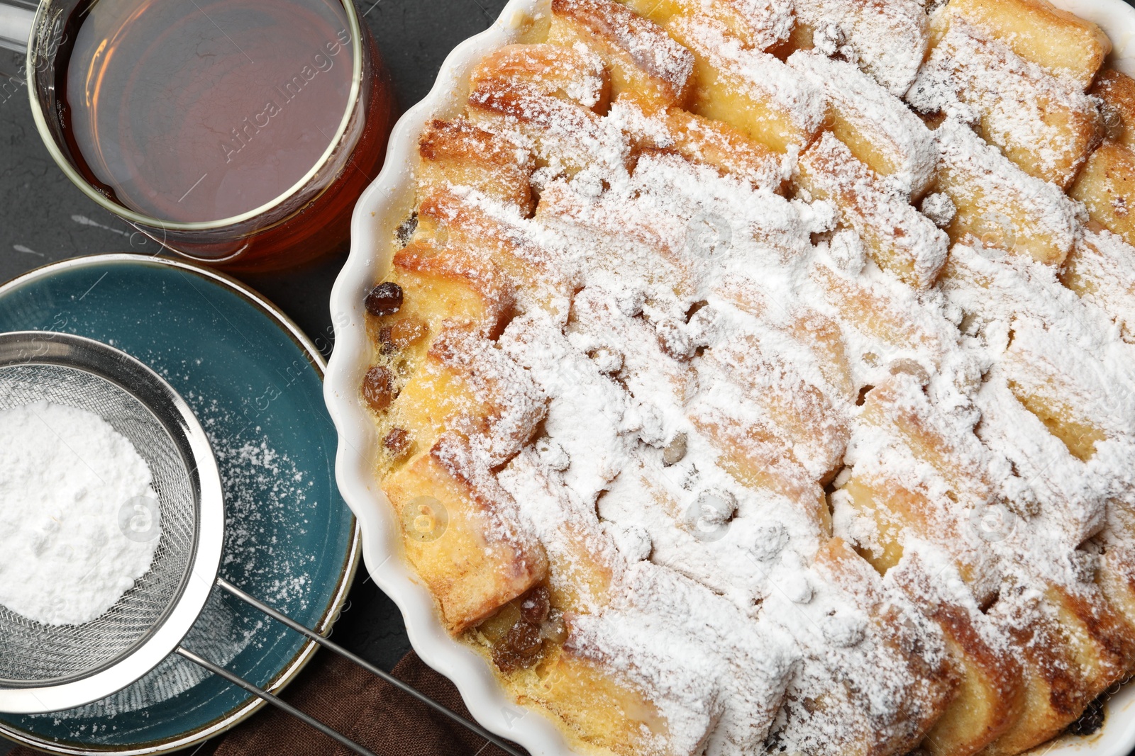 Photo of Delicious bread pudding with raisins, powdered sugar and tea on black table, flat lay