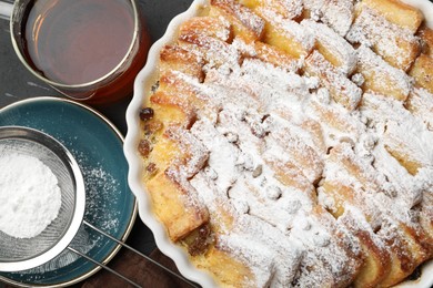 Photo of Delicious bread pudding with raisins, powdered sugar and tea on black table, flat lay