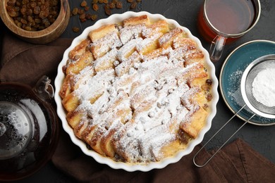 Photo of Delicious bread pudding with raisins, powdered sugar and tea on black table, flat lay