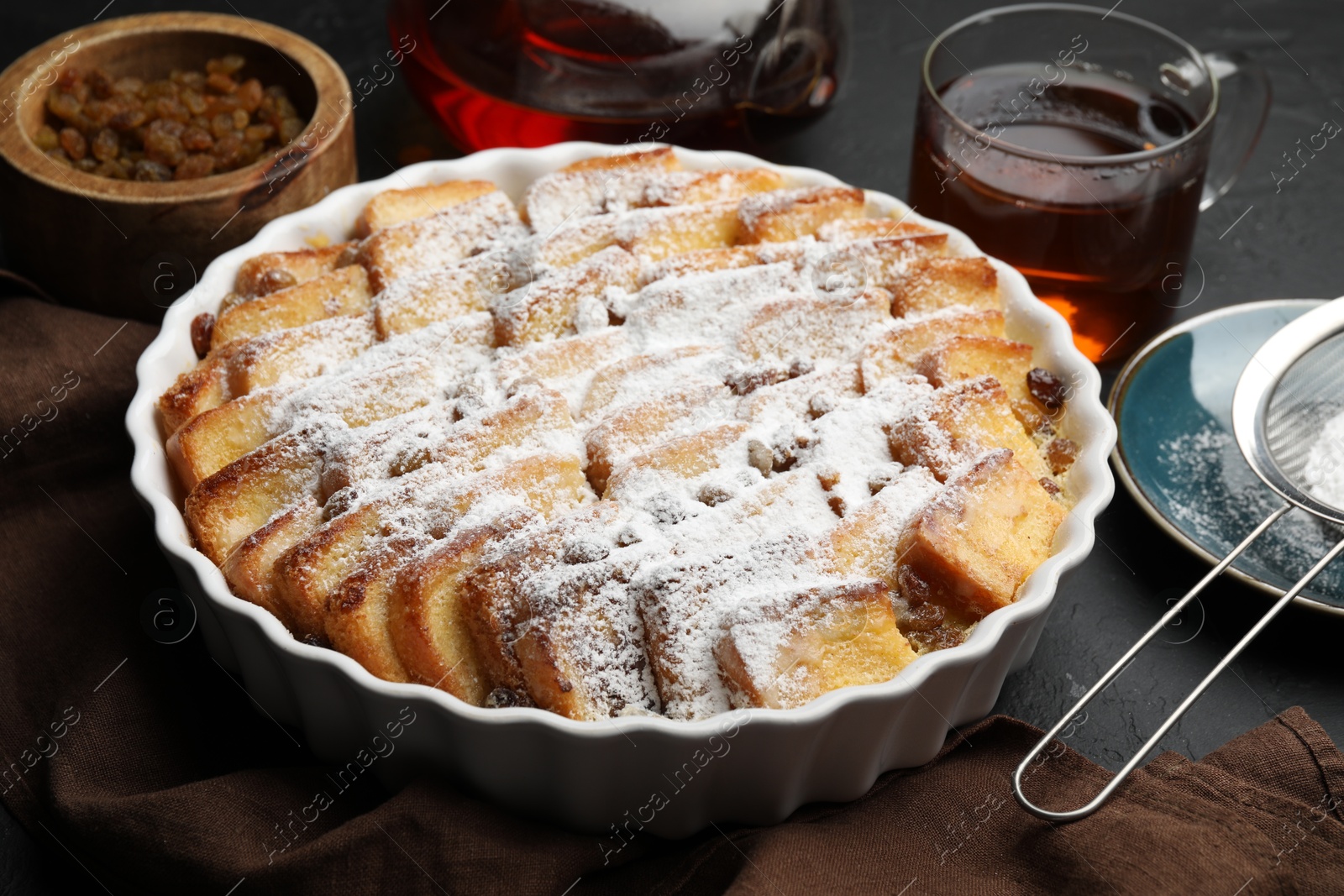 Photo of Delicious bread pudding with raisins, powdered sugar and tea on black table, closeup