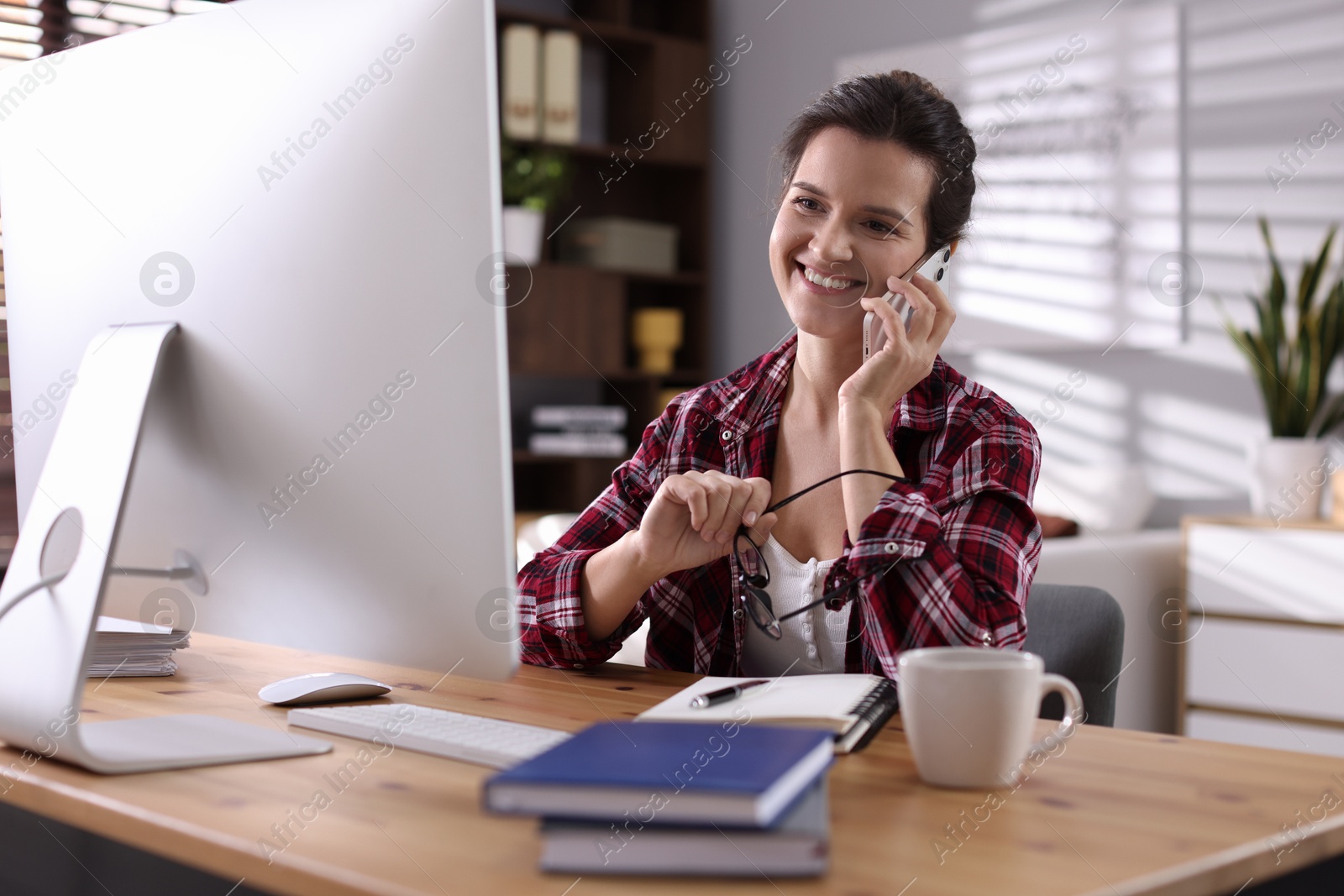 Photo of Happy woman talking by smartphone at desk with computer indoors. Home office