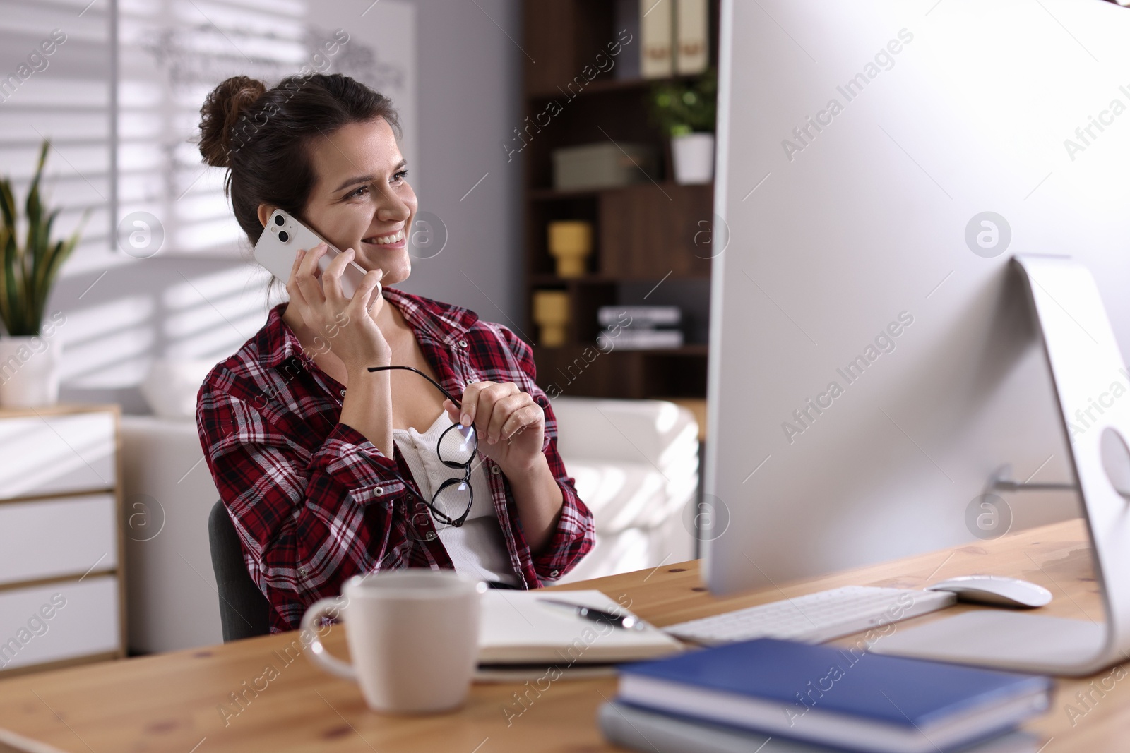 Photo of Happy woman talking by smartphone at desk with computer indoors. Home office