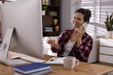 Happy woman talking by smartphone at desk with computer indoors. Home office