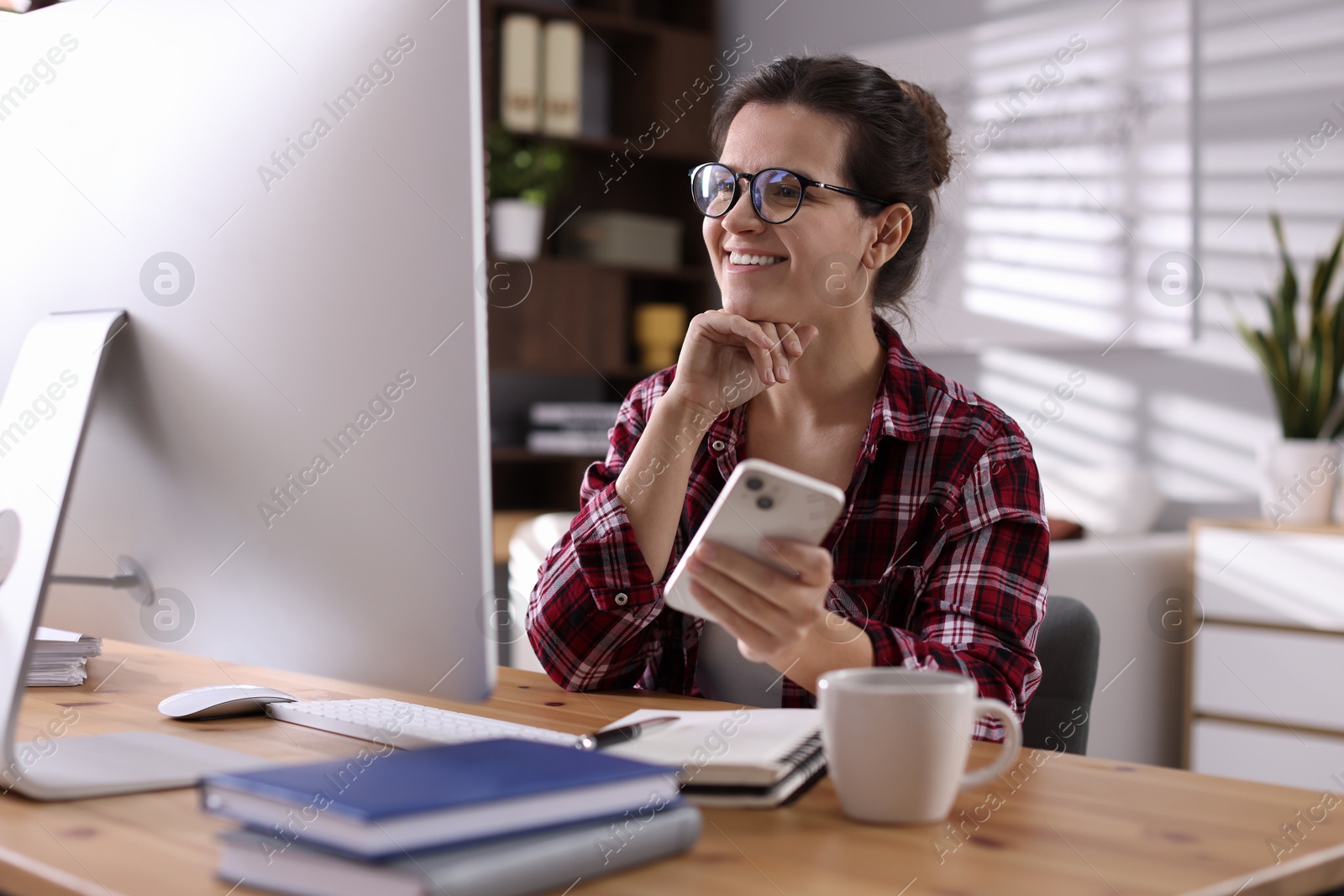 Photo of Happy woman holding smartphone and working with computer at desk indoors. Home office